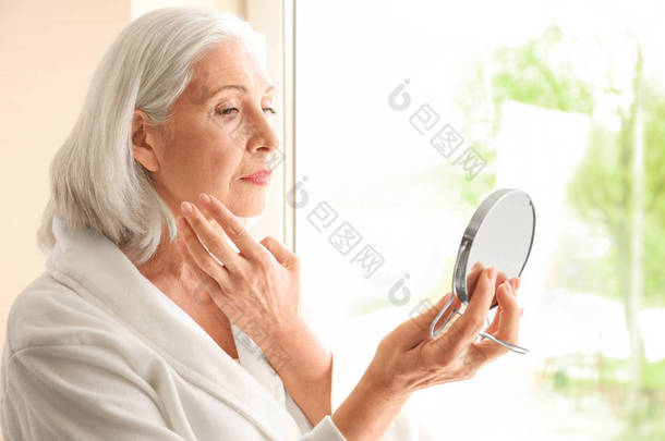 美丽的老妇拿着镜子和应用在家里面霜