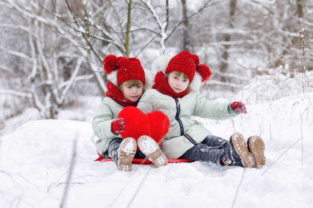 有趣的<strong>姐妹</strong>双胞胎坐在雪地上, 在冬季公园散步时, 手里拿着一颗巨大的红心.