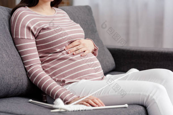 在沙发上坐着针织针和纱的年轻孕妇的裁剪照片