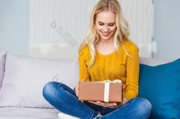 美丽微笑的年轻妇女拿着礼物盒, 而坐在沙发上在家