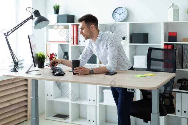一个年轻人站在办公室的桌子旁, 拿着一支铅笔和一杯咖啡。一个年轻人工作的文件和计算机.