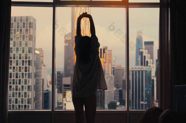 年轻修长的女人的剪影站在大窗口, 伸出手, 早上醒来在日出