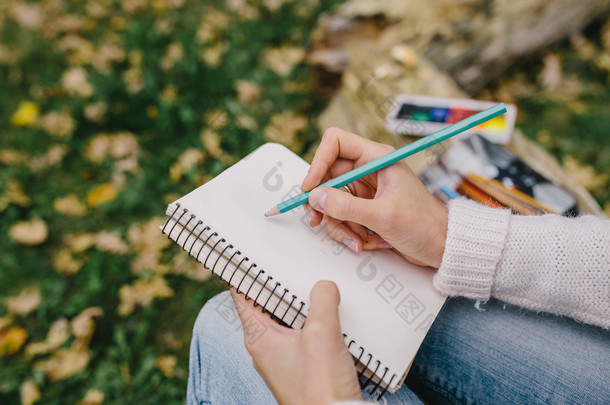 在草和落叶背景的笔记本上绘制彩色铅笔的剪画图像。特写女人在户外的笔记本上写