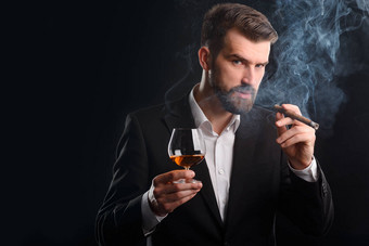 抽烟的男人喝酒。穿着正式服装的有吸引力的商人的横向肖像。一杯白兰地和一支烟熏雪茄在他手中.图片
