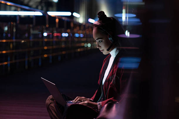 有吸引力的亚洲女孩在<strong>和服</strong>坐, 并使用笔记本电脑与耳机与霓虹灯在街上, 未来概念的城市