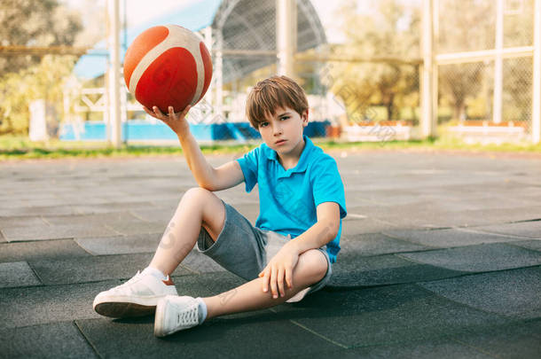 一个穿着蓝色T恤的漂亮男孩篮球运动员手拿着球坐在篮球场上。那位运动员在训练后正在休息.