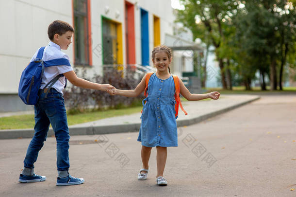 一个笑容满面的小女孩和一个背着背包的弟弟手牵着手，玩耍着，在教学楼附近玩得很开心