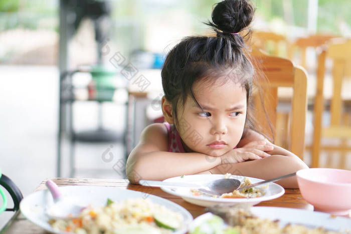 亚洲小孩可爱或小孩厌食症或伤心无聊的食物，或困倦的空余食物，撑着身子或面颊靠在木桌上吃早餐或午餐