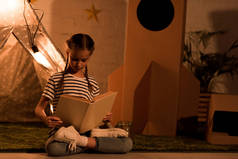 青春期前的孩子坐在莲花的姿势和阅读书籍在黑暗的房间