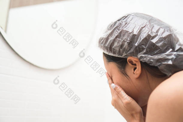 年轻的亚洲妇女在浴室洗澡前洗脸。美丽的女孩穿淋浴帽清洁脸上的浴室水槽和镜子。护肤及治疗, 晨生.