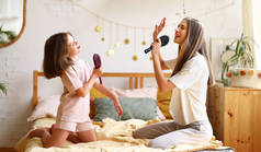 两个快乐的女孩假装是有名的歌手，坐在床上，用梳子像麦克风一样唱歌，让小姐姐和姐姐喜出望外。年轻的妈妈和可爱的小女儿在家里玩的开心