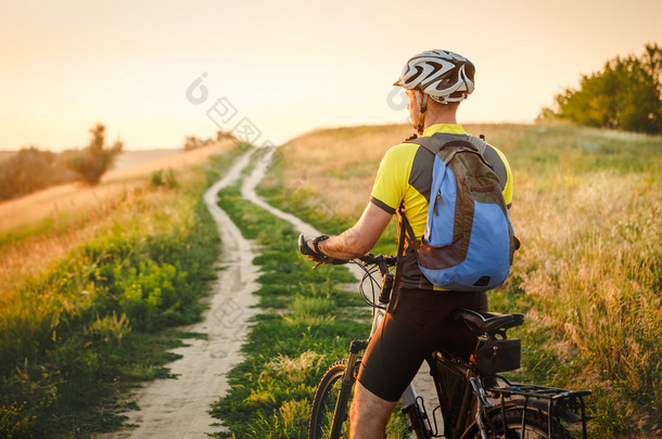 年轻男子在一条乡村公路穿过夕阳夏天草地上骑自行车