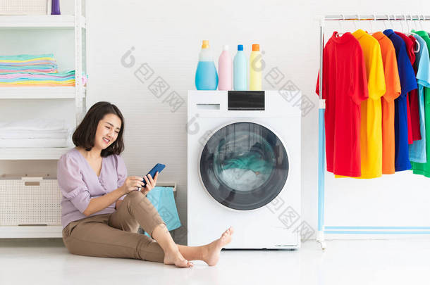 在等待洗衣机洗衣服的过程中，坐在地板上看着智能手机的亚洲女人带着一种放松的心情在洗衣房里笑着