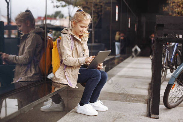 放学后，一个笑着可爱的女孩坐在玻璃楼旁边的台阶上等公共汽车，在平板电脑上<strong>学习</strong>，并通过视频进行<strong>交流</strong>.