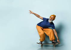 留着胡子的老人穿着T恤，太阳镜，橙色裤子，帽子，胶鞋。骑黑色滑板，在蓝色背景上摆姿势