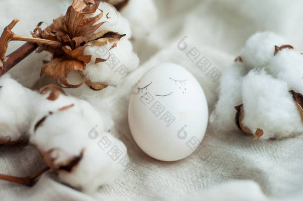 有脸的白色复活节彩蛋。旁边是一条灰色亚麻布背景的棉花枝条