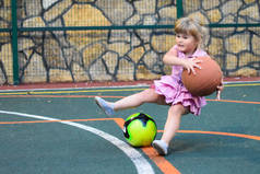一个小女孩带着球倒在运动场上。一个拿着大篮球的孩子