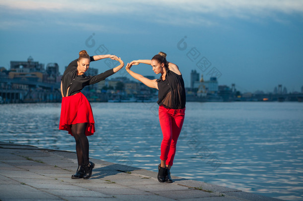 在河附近城市的背景下，两个年轻漂亮的双胞胎姐妹在跳舞 waacking 舞蹈。在夏天时间上显示不同的风格和现代舞蹈与黑色和红色的衣服，近水的姿势.