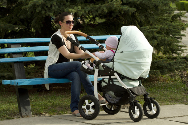 母亲坐在长凳上，她的女儿在婴儿推车.