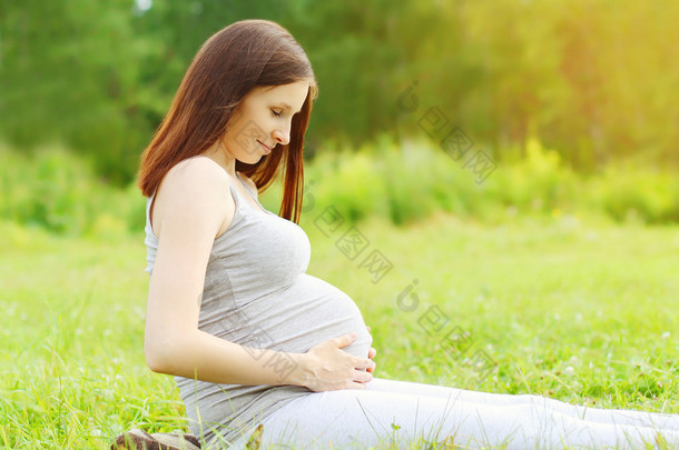 Portrait of <strong>happy</strong> pregnant woman sitting on the grass enjoys sun
