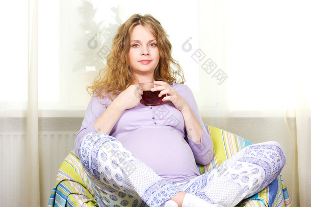 怀孕的金发女郎坐在家里喝茶。Portrait.Beautiful 怀孕卷曲金发
