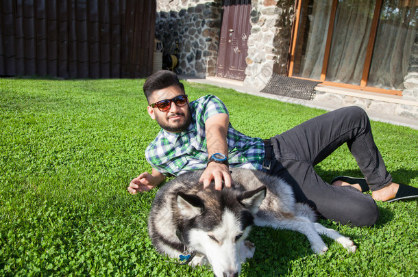 胡子和时尚发型的一个时尚中间东部人是休息和享受美丽的绿色草地上一天时间和 Hasky 狗跟他玩。看着相机微笑着幸福.