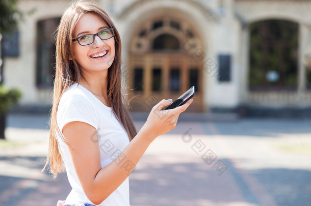 Cheerful young female student is using telephone outdoors