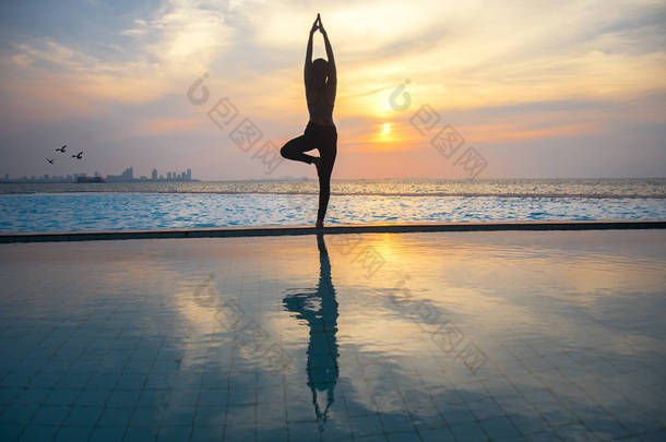 剪影年轻女人上练瑜伽的游泳池和海滩日落时分