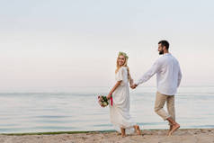 快乐的新娘和新郎牵手走在沙滩上的婚礼花束