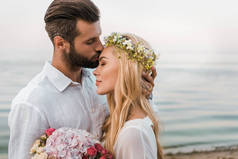 侧面观看英俊的新郎亲吻迷人的新娘额头在海滩上