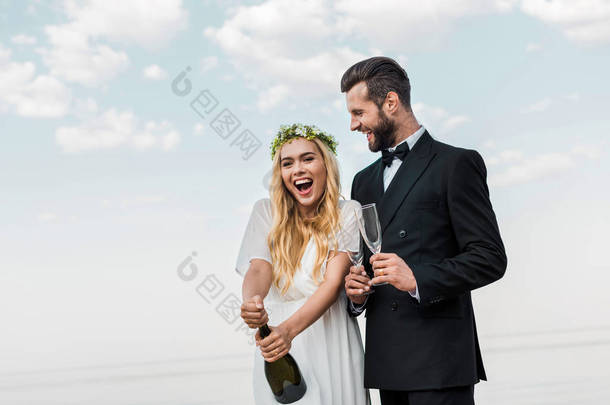 快乐的新娘在白色礼服打开香槟瓶在海滩上