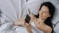 美丽的布鲁内特的肖像使用触摸屏智能手机在她的床上,与朋友聊天,使用社交媒体,张贴图片,分享想法。自上而下拍摄.