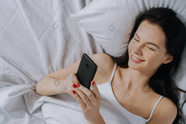美丽的布鲁内特的肖像使用触摸屏智能<strong>手机</strong>在她的床上,与朋友<strong>聊天</strong>,使用社交媒体,张贴<strong>图片</strong>,分享想法。自上而下拍摄.