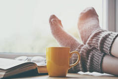 妇女休息保持腿在温暖的袜子桌与早晨咖啡和读书书