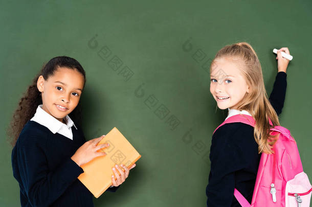 <strong>非洲</strong>美国女学生拿着书附近的同学与一块粉笔附近的绿色黑板