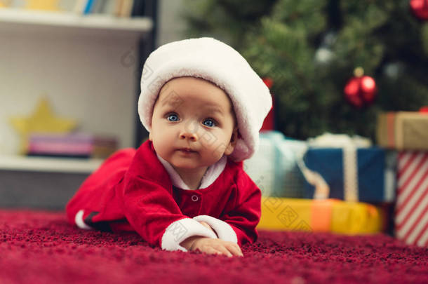 美丽的小婴儿的特写肖像躺在圣诞树前的红色地毯和礼物的圣诞老人西装