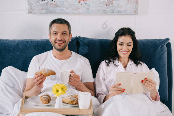 快乐的女人读书附近欢快的丈夫拿着羊角面包和杯子在床上 