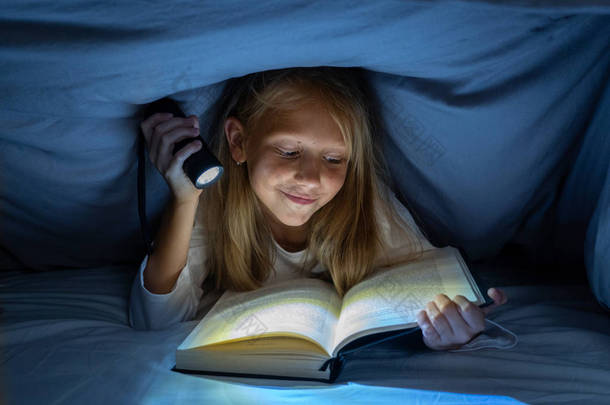 快乐聪明的女孩读童话般的书躺在黑暗中的被套床下拿着一盏灯笼在阅读技能文学学校的成功和教育的概念.