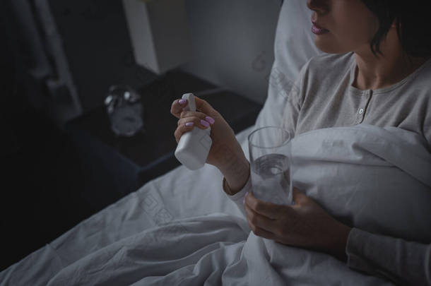 睡眠障碍妇女在卧室拿着一杯水和装有安眠药的瓶子的剪影 