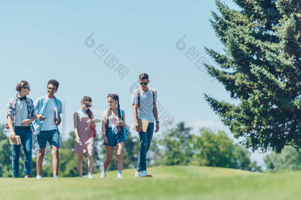 多民族青少年学生在公园散步时用书本和滑板交谈  