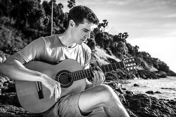 帅气的微笑吉他手玩音乐选址上海滩岩。黑色白色户外写真.