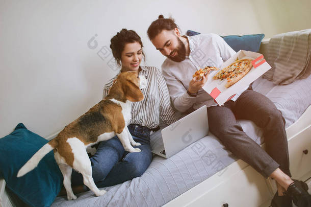 年轻的时髦夫妇家中吃披萨笔记本电脑与狗