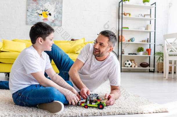 微笑的父亲和儿子坐在地毯上，在家里玩玩具车