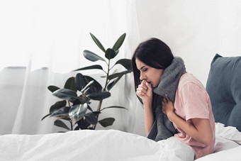 生病的年轻妇女坐在床上咳嗽, 而痛苦喉咙痛图片