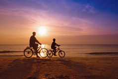 父亲和儿子在海滩上落日下，骑自行车的人的家庭剪影 