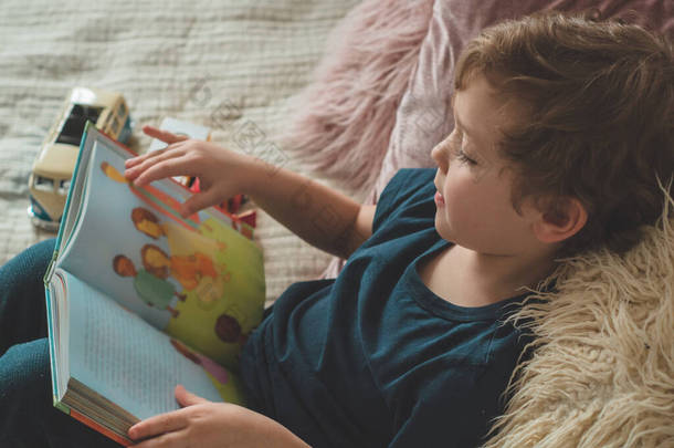 A little boy sits on a bed with your toys in living room watching pictures in story book