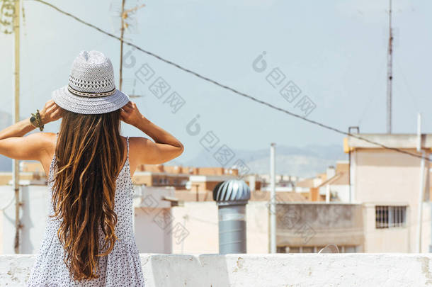 长头发的女人戴着<strong>帽子</strong>看着地平线上的城市, 从后面, 穿着在一个夏天的礼服