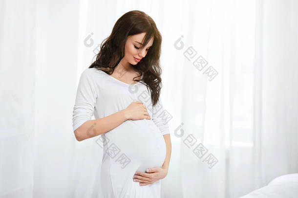 怀孕妇女与婴孩腹部在室内。美丽的微笑的女孩在白色的内饰。高分辨率