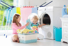 儿童在洗衣房内设有洗衣机或滚筒烘干机。孩子们帮助做家事。现代家庭设备和洗涤洗涤剂在白色阳光的家。烘干机架清洗洗衣服. 