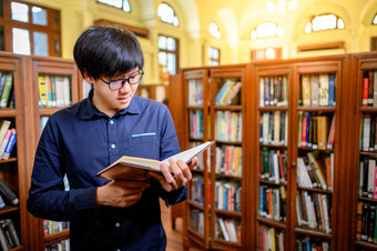 聪明的亚洲人大学生戴着眼镜，坐在老式书架上看书。高校图书馆的教材资源用于教育科目和研究.教育机会奖学金.图片